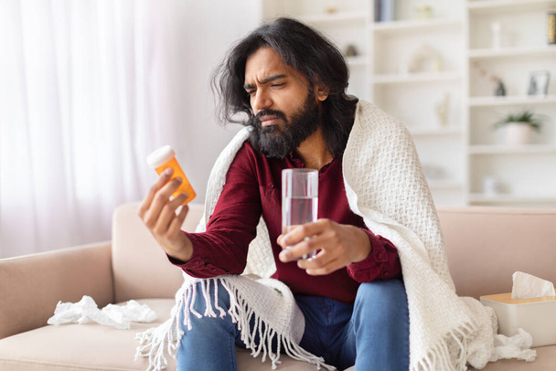 Un uomo indiano preoccupato avvolto in una coperta guarda confusamente una bottiglia di pillole mentre tiene in mano un bicchiere d'acqua, illustrando malattia e trattamento. - Foto, immagini