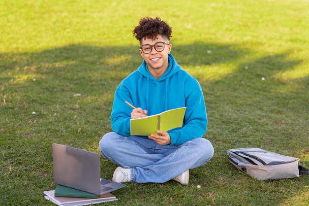 Un giovane brasiliano che indossa occhiali da vista scrive in un quaderno mentre è seduto sull'erba con laptop e zaino - Foto, immagini