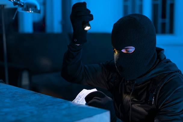 Um ladrão focado usa uma lanterna para roubar dinheiro em um quarto escuro, possivelmente procurando informações pessoais - Foto, Imagem
