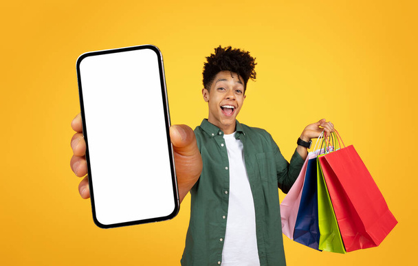 Naurava nuori musta mies ojentaa älypuhelimen kanssa mockup näytön ja ostoskassit - Valokuva, kuva