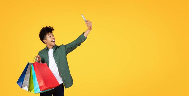 Energetischer junger Schwarzer, der einen Selfie-Moment mit lebendigen Einkaufstüten in der Hand festhält - Foto, Bild