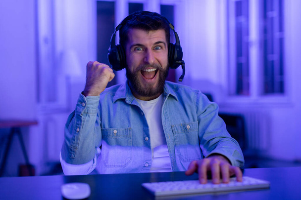 Podekscytowany gracz facet w słuchawki gesty wygranej podczas gry online oświetlone przez ekran komputera - Zdjęcie, obraz