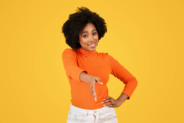Donna sorridente con capelli ricci in dolcevita arancione che offre una stretta di mano, raffigurante un gesto amichevole e professionale, abbinato a pantaloni bianchi, su uno sfondo giallo allegro - Foto, immagini