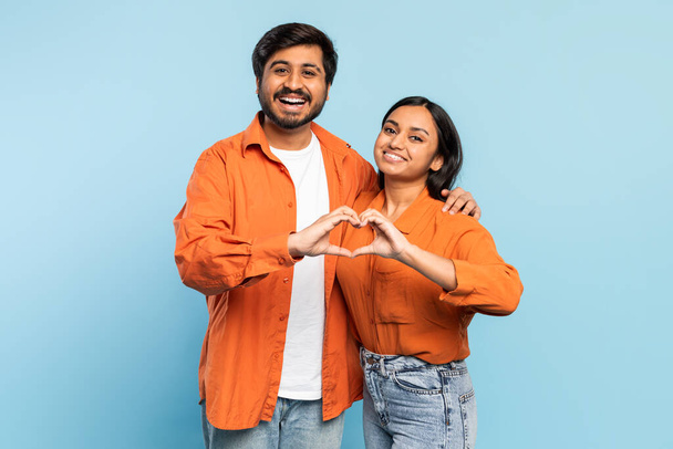 Smiling indian couple forms a heart shape with hands, symbolizing love and connection against a blue background - Photo, Image