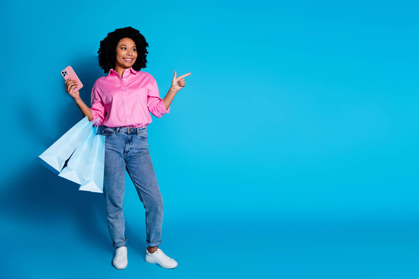 Full length portrait of pretty young woman hold smart phone bags direct finger empty space wear pink shirt isolated on blue color background. - 写真・画像