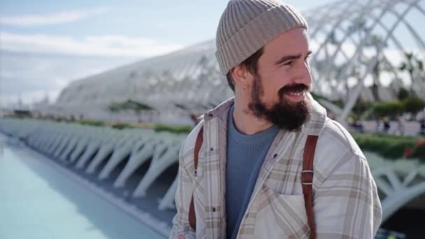Genç, gülümseyen, beyaz, çekici bir adam cep telefonuyla yazarken arkasına bakıyor ve yürüyor. Güneşli bir kış gününde Valencia 'daki bilim parkında mutlu bir erkek turist. - Video, Çekim