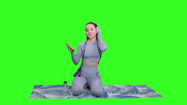 Smiling sporty woman listening the music by headphones while holding phone and sitting on yoga mat on green isolated background. Fitness, sport, healthy concept - Footage, Video