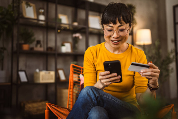Japanische reife Frau mit Brille sitzt auf Stuhl mit Kreditkarte und Handy Online-Shopping zu Hause - Foto, Bild