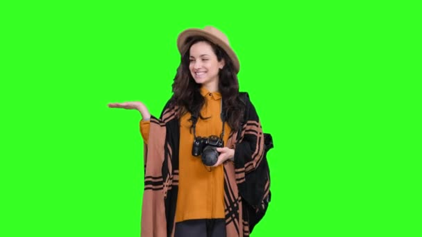 Smiling young female tourist in stylish hat using camera while pointing copy space by hand on green isolated background. Trip, tourism, adventures concept - Footage, Video