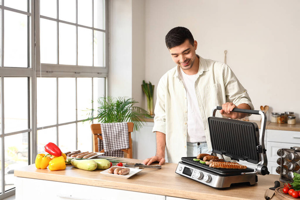 Nuori mies ruoanlaitto maukkaita makkaroita ja vihanneksia moderni sähkögrilli pöydässä keittiössä - Valokuva, kuva