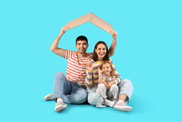 Familia feliz con cartón en forma de techo soñando con su nueva casa sobre fondo azul - Foto, Imagen