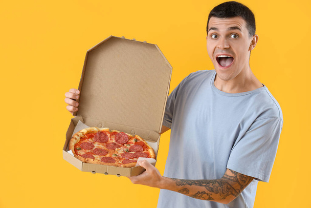 Zaskoczony młody człowiek ze smaczną pizzą pepperoni na żółtym tle - Zdjęcie, obraz