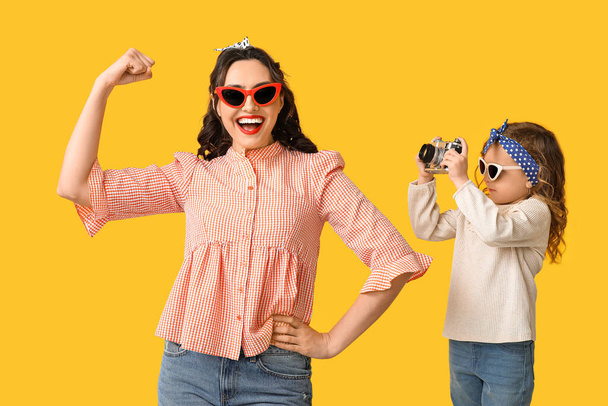 Schöne Pin-up-Frau zeigt Muskeln und ihre Tochter mit Fotokamera auf gelbem Hintergrund - Foto, Bild