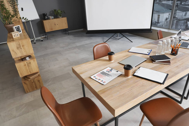 В современном конференц-зале подготовлены деревянный стол, кресла и канцелярские принадлежности для деловой встречи - Фото, изображение