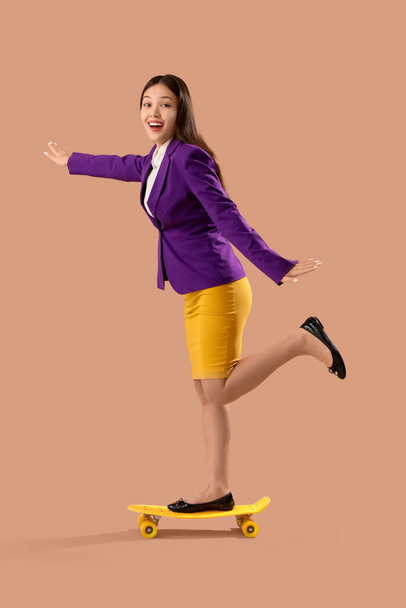 Смішна шокована азіатська бізнес-леді верхи скейтборд на коричневому фоні - Фото, зображення