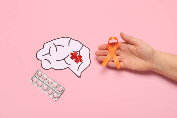 Kobieca ręka z pomarańczową wstążką, papierowy mózg, kawałek układanki i pigułki na różowym tle. Miesiąc świadomości stwardnienia rozsianego - Zdjęcie, obraz