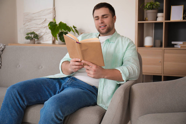 Junger Mann liest Buch auf grauem Sofa zu Hause - Foto, Bild