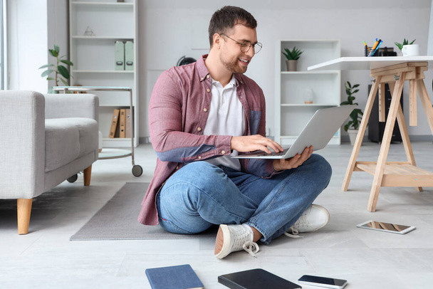 Programmatore maschio con computer portatile e diversi gadget seduti sul pavimento in ufficio - Foto, immagini