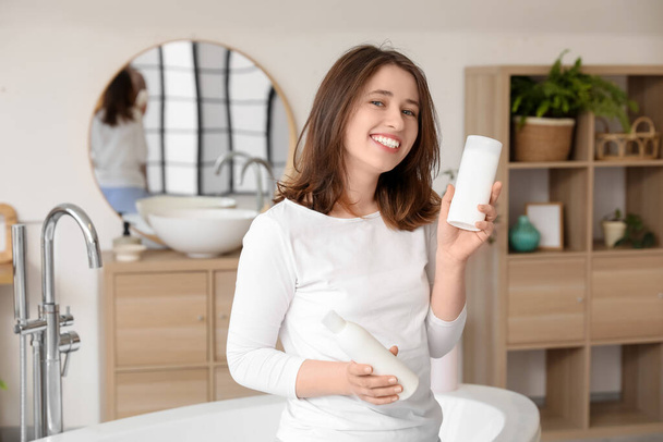 Красива молода щаслива жінка з пляшками продуктів для волосся у ванній - Фото, зображення