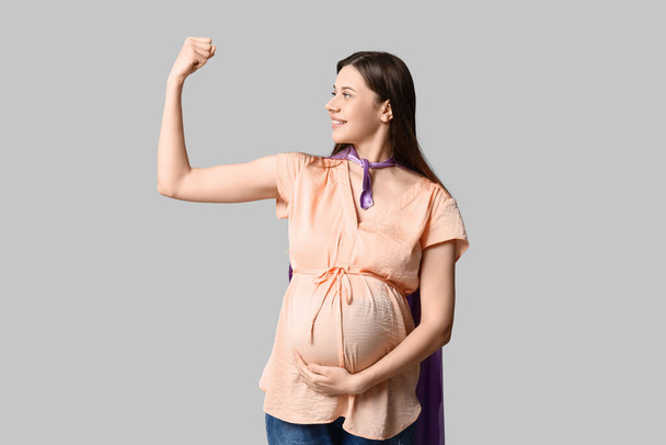 Mooie jonge zwangere vrouw in superheld cape tonen spieren op grijze achtergrond - Foto, afbeelding
