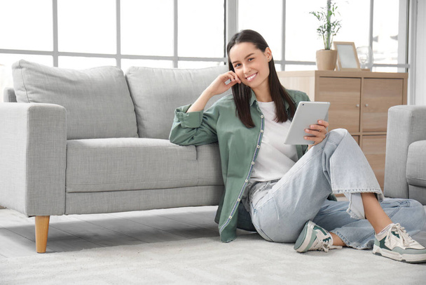 Красива молода жінка з планшетним комп'ютером, що сидить на підлозі біля сірого дивана у вітальні - Фото, зображення
