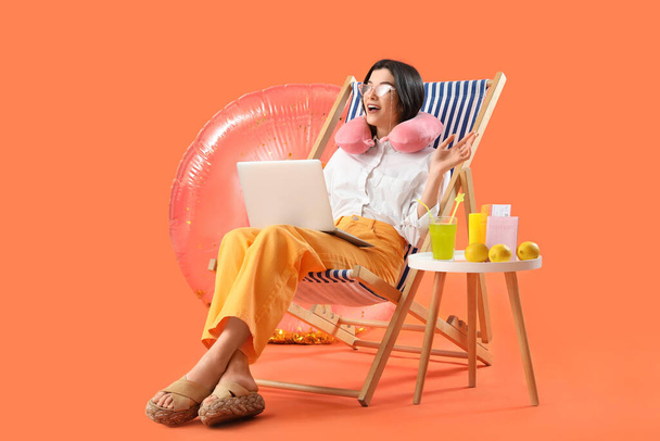 オレンジ色の背景で夏休みの準備ができているラップトップとビーチアクセサリーを持つ若いビジネスマン - 写真・画像