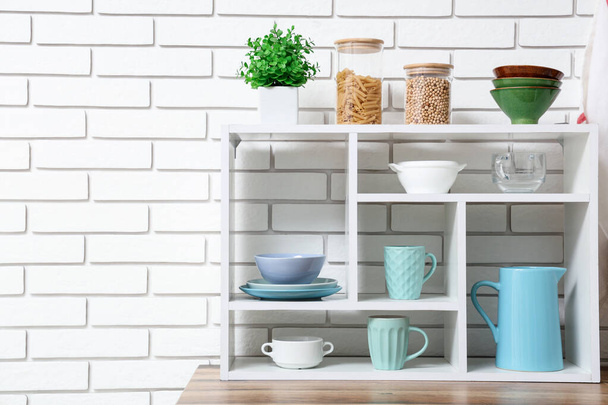 Shelving unit with set of clean tableware and cups near white brick wall - Photo, Image