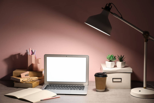Local de trabalho com laptop moderno, xícara de café, livros e lâmpada brilhante perto da parede rosa - Foto, Imagem