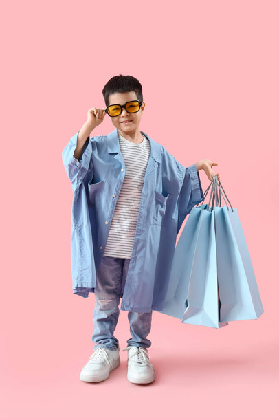 Leuke kleine Aziatische jongen in volwassen kleding met boodschappentassen op roze achtergrond - Foto, afbeelding