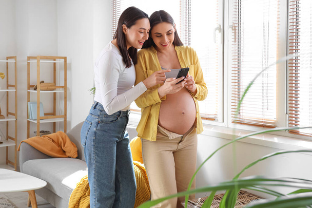 Fiatal terhes leszbikus pár mobiltelefonnal otthon - Fotó, kép