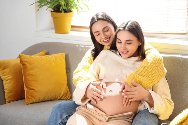 Young pregnant lesbian couple hugging on sofa at home - Foto, afbeelding