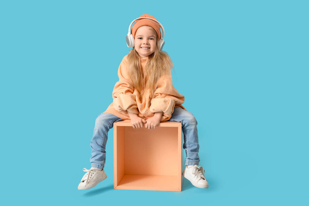 Cute little girl in adult clothes with headphones sitting on blue background - Photo, Image