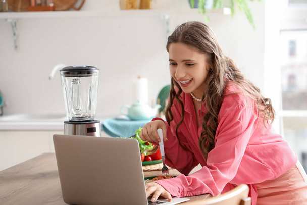 Νεαρή γυναίκα με φορητό υπολογιστή στο τραπέζι τρώει νόστιμα ζυμαρικά στην κουζίνα - Φωτογραφία, εικόνα