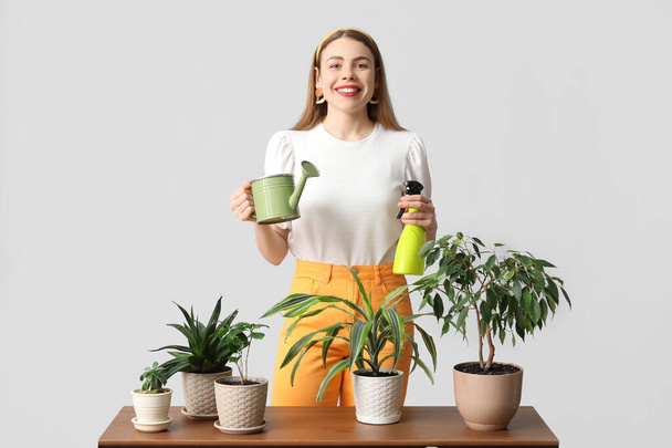 Красива молода жінка з лійкою, розпилювач води і кімнатні рослини на столі на сірому фоні - Фото, зображення