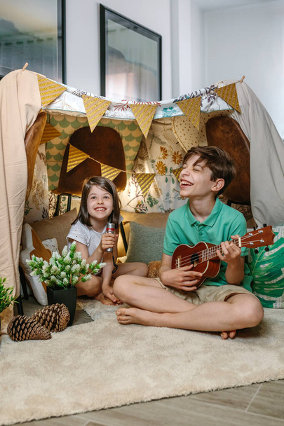 Ukulele çalan ve oturma odasındaki el yapımı çadırda şarkı söyleyen mutlu çocuklar. Kız ve erkek evdeki sıcak çadırda gülüyorlar. Ev kavramında konaklama ve tatil kampı. - Fotoğraf, Görsel