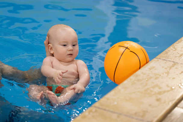 Baby versucht, den Ball im Schwimmbad zu fangen, schaut aber auf etwas anderes. Wasserspiele für Neugeborene - Foto, Bild
