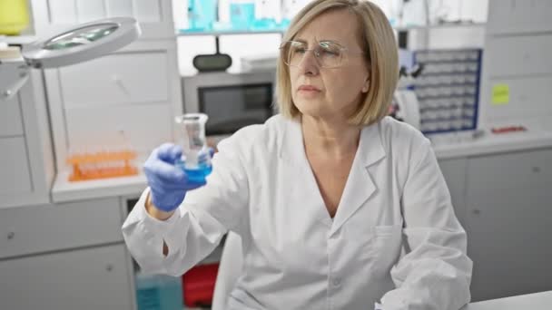 Kypsä naistutkija laboratoriossa tutkimassa pulloa, jossa on sinistä nestettä, kuvaten ammattimaisuutta ja keskittymistä - Materiaali, video