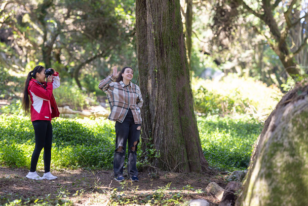 Zwei Freundinnen stehen nebeneinander neben einem hohen Baum im Freien. Beide Frauen sind leger gekleidet und scheinen in Gespräche verwickelt zu sein oder die Umgebung zu genießen. - Foto, Bild