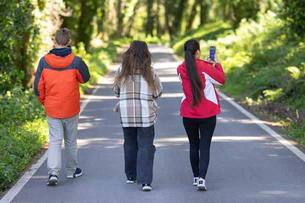 A group of friends walking along a road, moving in a straight line. They are wearing casual clothing and appear to be engaged in conversation while enjoying the outdoors together. - Photo, Image