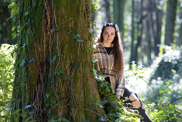 Een vrouw zit naast een boom in een dicht bos, omgeven door weelderig groen. Ze lijkt ontspannen, genietend van de vredige sfeer terwijl ze buiten tijd doorbrengt met vrienden.. - Foto, afbeelding