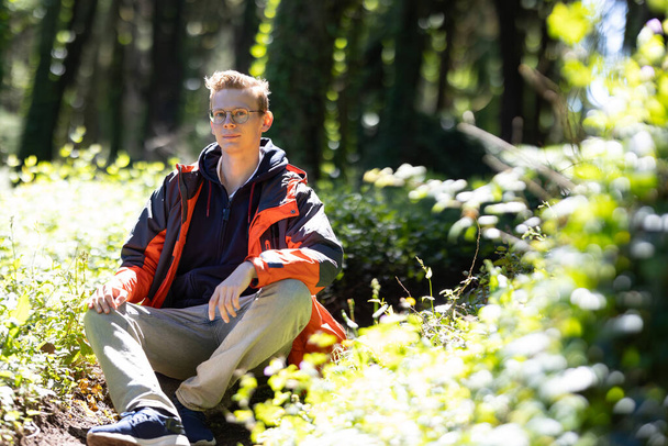 Mladý chlapec sedí na velké skále mezi stromy v zalesněné oblasti. Vypadá uvolněně a zamyšleně, zaujímá své okolí. - Fotografie, Obrázek