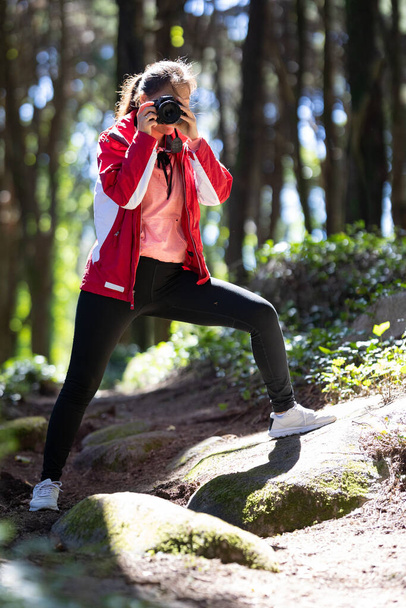 Uma mulher de pé na floresta, segurando um telefone da câmera para tirar uma selfie de si mesma. O fundo mostra árvores e folhagens típicas de um cenário florestal. - Foto, Imagem