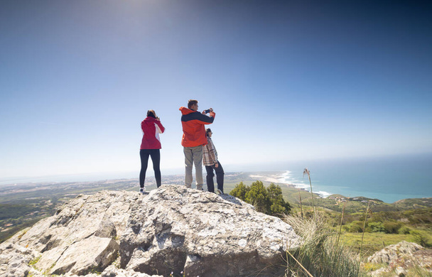 Three friends are standing together on the peak of a mountain, gazing out at the vast expanse of the ocean spread out before them. The trio appears to be enjoying the panoramic view of the sea. - Photo, Image