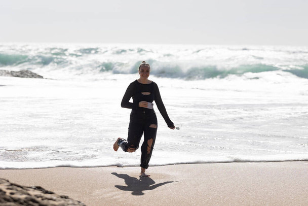 Märkäpukuinen nainen juoksee rannalla. Hänen hiuksensa lentävät hänen takanaan, kun hän liikkuu energisesti hiekkarannalla ystävien kanssa.. - Valokuva, kuva