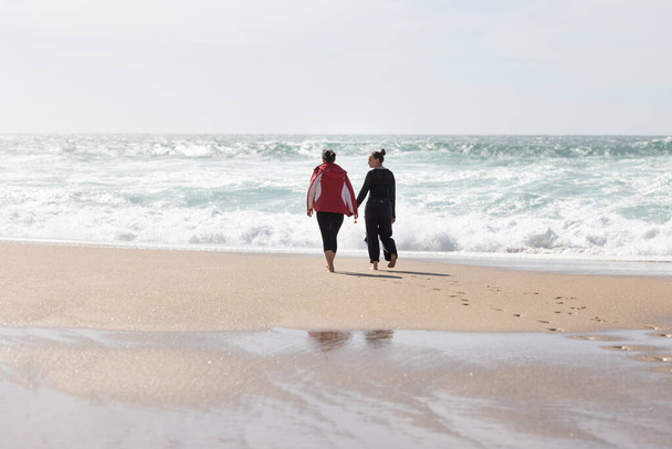 Para osobników, prawdopodobnie przyjaciół, spacerująca po piaszczystej plaży, trzymając się za ręce. Obie postacie są zaangażowane we wspólną działalność, ciesząc się nadmorską atmosferą. - Zdjęcie, obraz