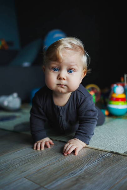 Un bebé de 7 meses se arrastra sobre una alfombra. El niño sostiene su cabeza. Un bebé aprende a arrastrarse por el suelo. Un niño en un mameluco gris. - Foto, imagen