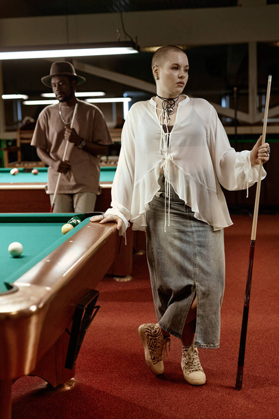 Vertical full length portrait of bald young woman standing by pool table in nightclub and holding cue stick - Photo, Image