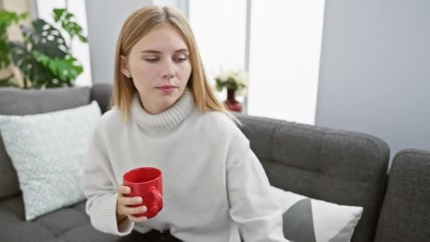 Mujer caucásica en un suéter blanco relajándose en un sofá gris con una taza roja y un teléfono inteligente en casa. - Imágenes, Vídeo