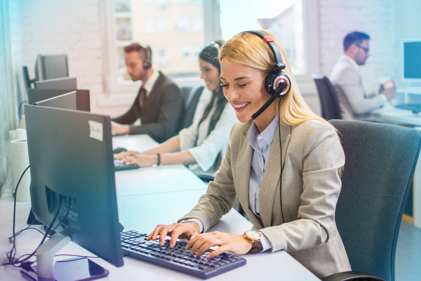 Young blonde haired woman in headset, sitting in office, working as operator of call center or support service with her colleagues. - Photo, Image