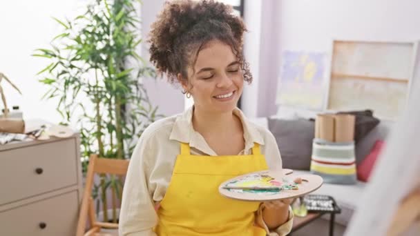 Cheerful hispanic artist woman with curly hair flaunts dazzling dental health, pointing at her teeth, happily sitting in art studio - Footage, Video